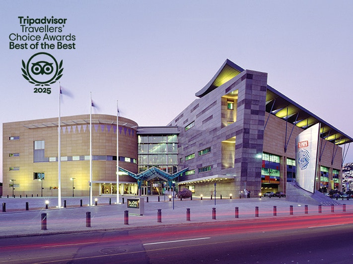 Te Papa at dusk. Photo by Te Papa Imaging. Te Papa (108024) A large building with strange angles and windows lit up. There is a road in the foreground where there are lone-exposure car light beams. There is a logo in the top left that says Tripadvisor Travellers Choice Awards Best of the Best and an image of an owl.