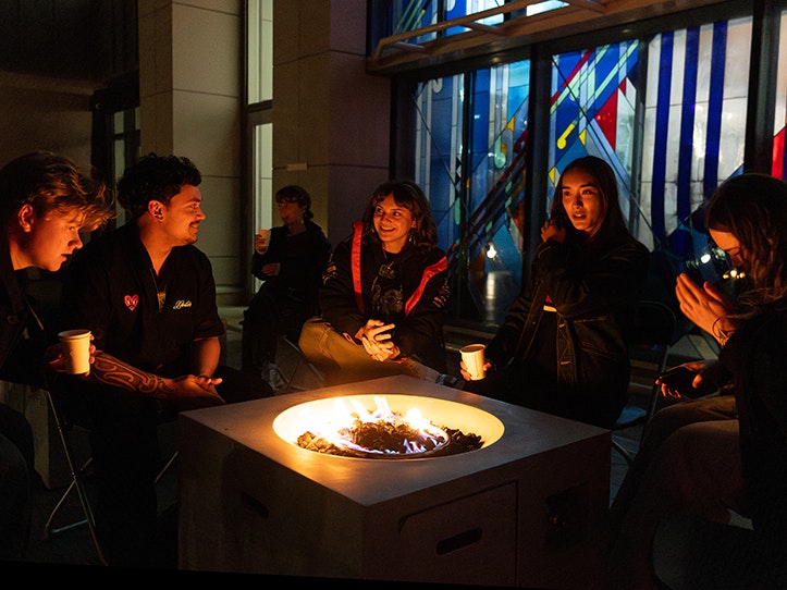 Matariki After Dark, 2025. Photo by Mel Waite. Te Papa (266475) Five people sitting around a fire pit beside a stained-glass window.