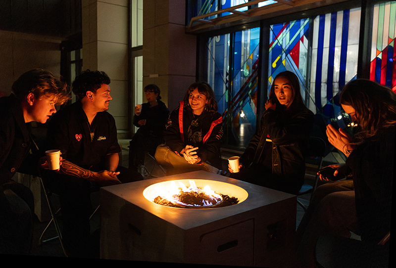 Five people sitting around a fire pit beside a stained-glass window.