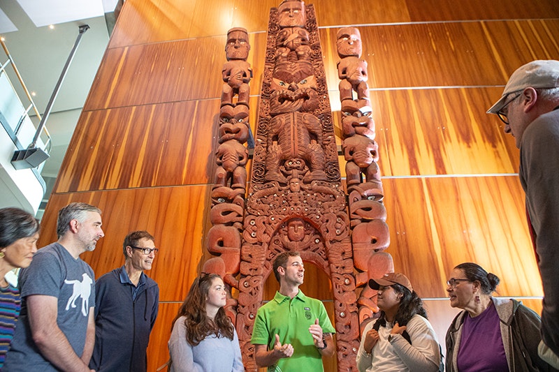 Several people are gathered at the base of a large waharoa carving in a museum. They are listening to the person in the middle.