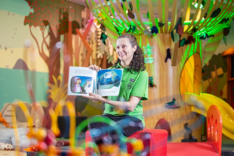A woman in a green shirt is holding a picture flat children's book up and reading to children off camera.
