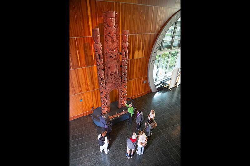A man in a green top is pointing at a very tall wooden carving. He is describing something to the man crouched next to two small children.