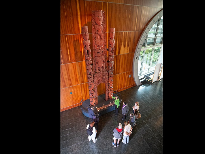 Visitors at Te Papa, 2025. Photo by Jeff McEwan. Te Papa (267137) A man in a green top is pointing at a very tall wooden carving. He is describing something to the man crouched next to two small children.