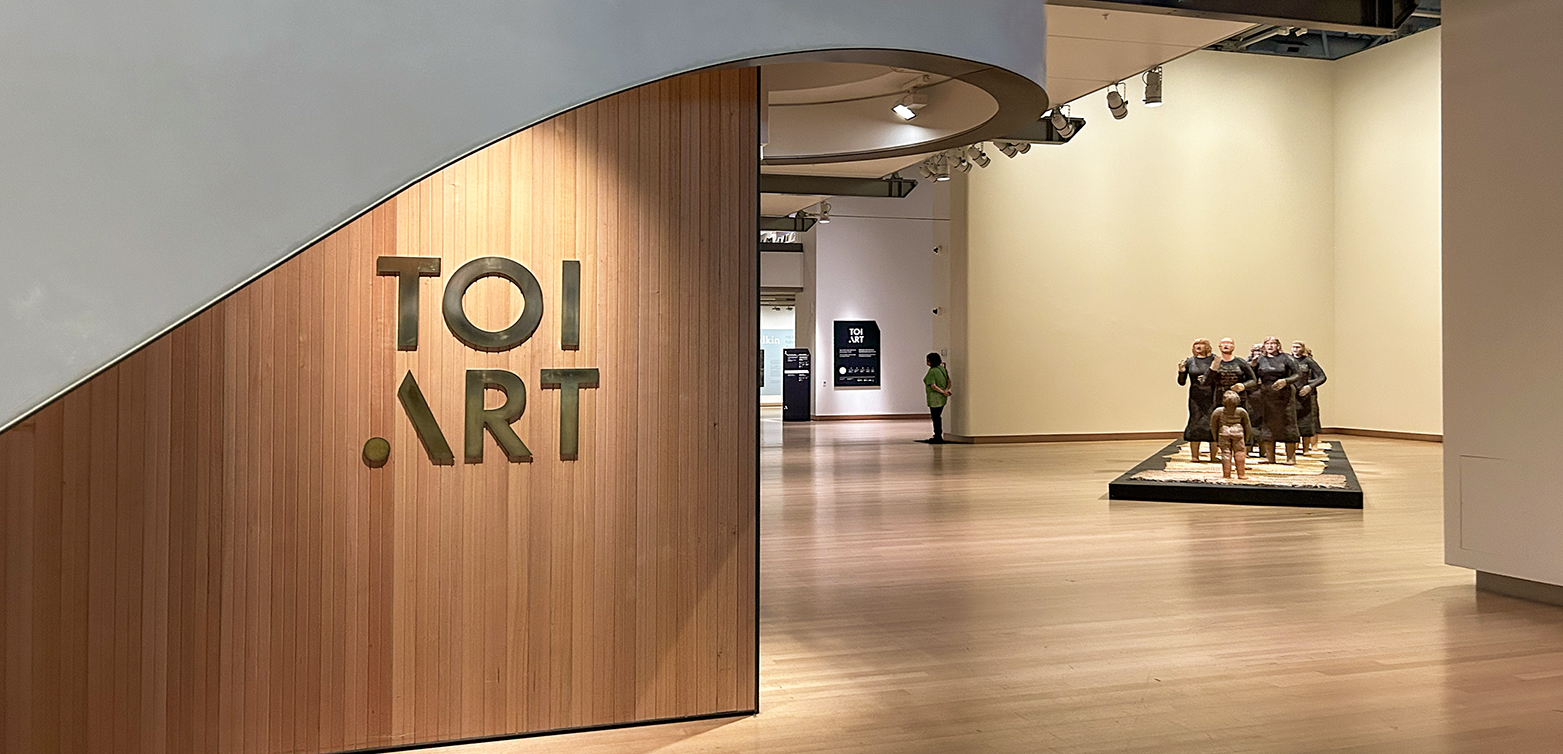 A gallery space with several life-size clay statues of people in a group in the mid-distance. There is a sign in the foreground that says Toi Art.