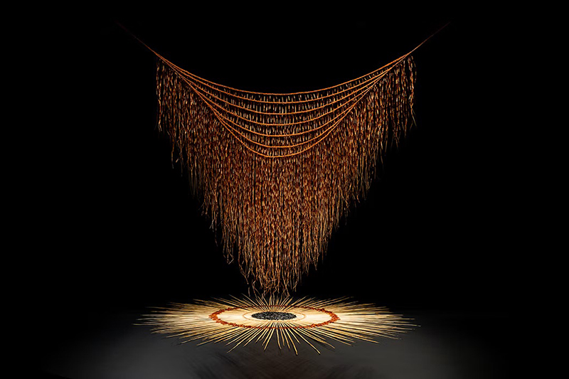 A black rectangle with an artwork of a woven cloak hanging over a circle of similar woven material.