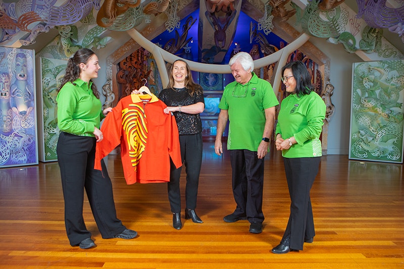 Three people in green shirts and one person is in a black shirt. Two of them are holding another shirt that is yellow and orange.