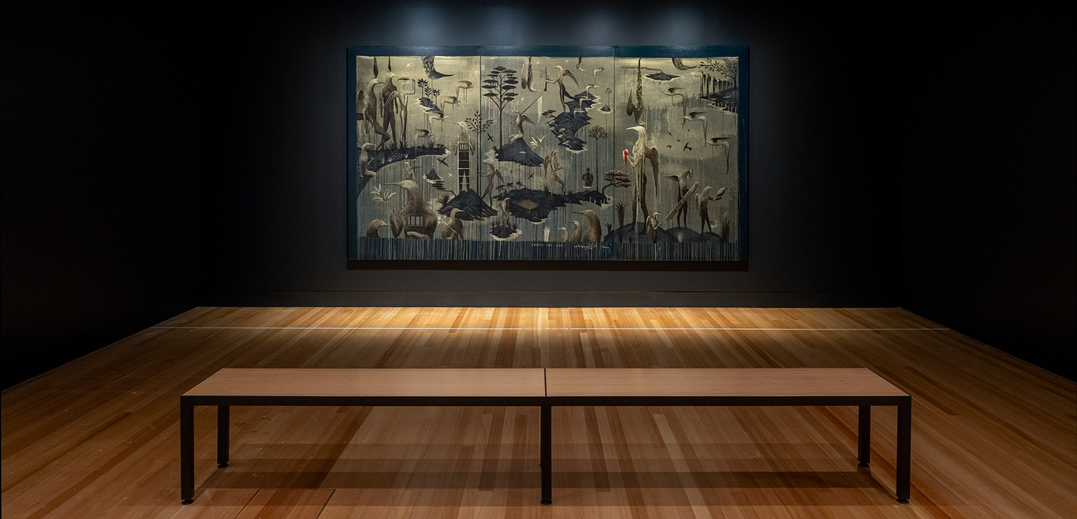 A dark room with a wooden floor and a wooden bench in front of a large rectangular artwork of bird men.