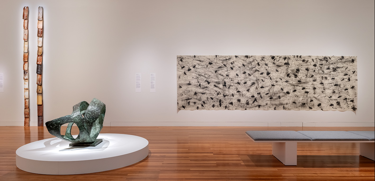 Installation view of three artworks, two on the wall and one sculpture on a round short plinth. There is also a long seat in shot.