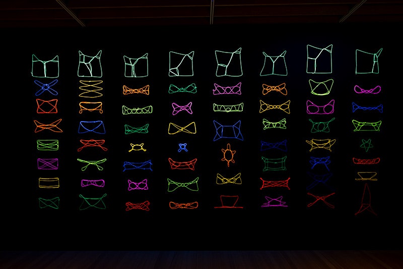 A black wall with eight columns of neon-coloured patterns created with string.