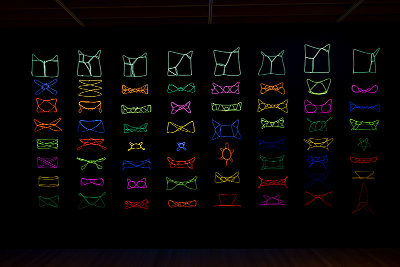 A black wall with eight columns of neon-coloured patterns created with string.