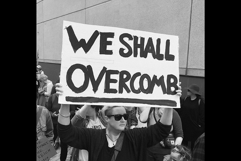 A black and white photo of a woman holding a large placard that says "We shall overcomb".