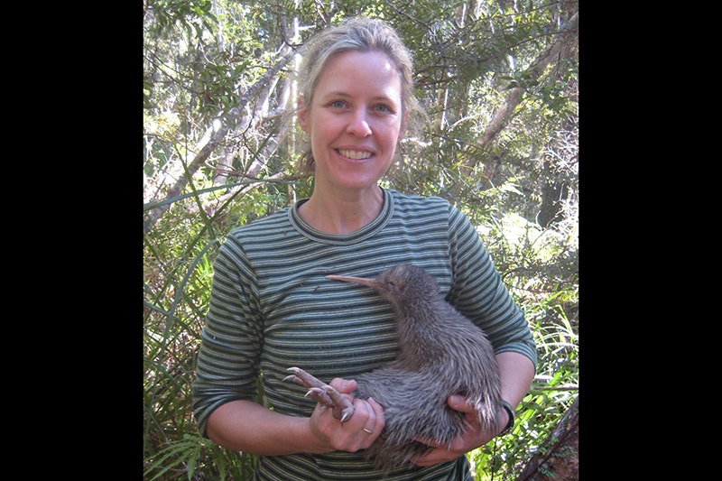 A woman in a striped top is standing in the forest and holding a live kiwi bird.