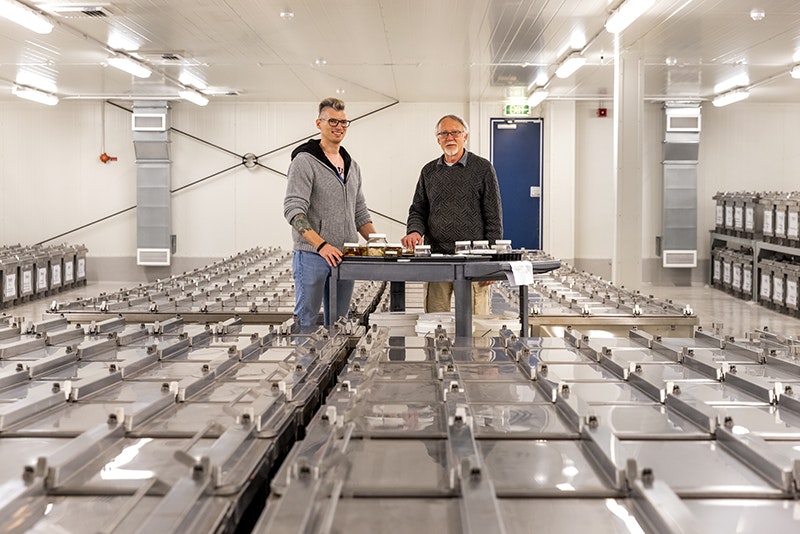 Two people are standing in the middle of a room with a trolley of fish specimens in jars in front of them. Surrounding them are large metal tanks full of fish you cannot see.