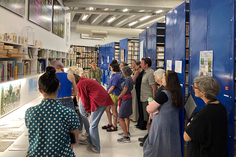 A group of people are standing in the botany collection store at Te Papa and looking at a desk with images of plants on them.