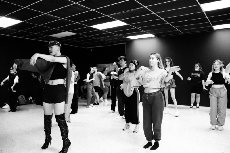 A black and white photograph of people in a dance studio. The lead person is wearing knee high boots and a black hat and is demonstrating a move.