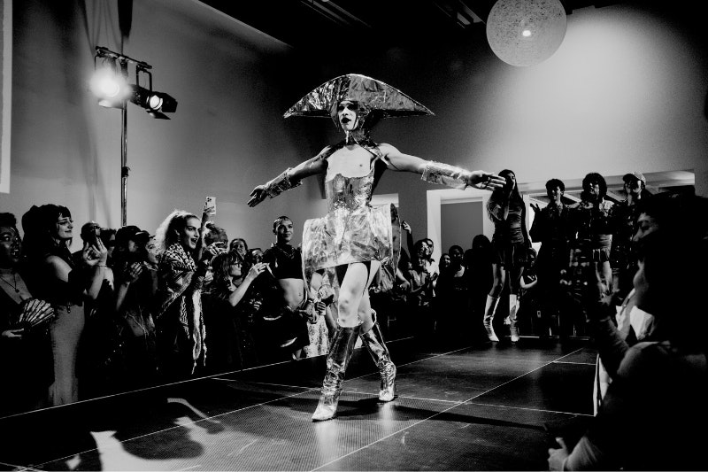 A person in a silver outfit is walking down a catwalk. The room is in a blue hue.