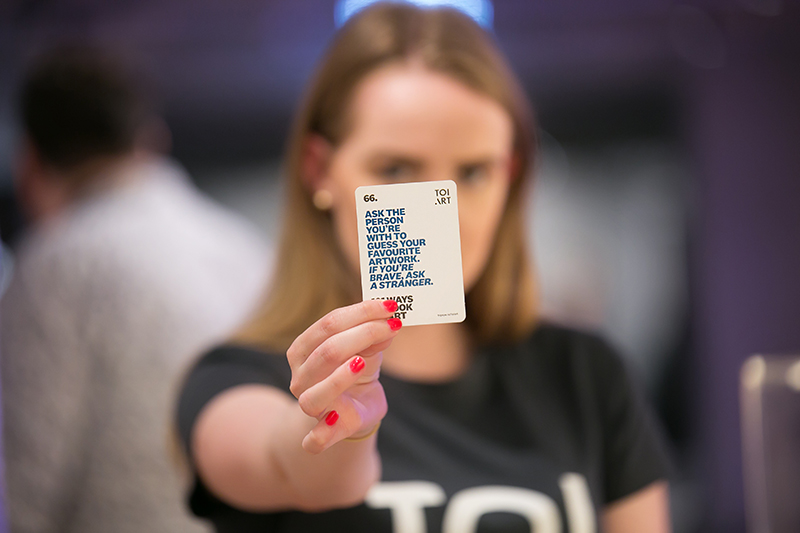 A woman in a black tshirt is holding up a small card that says "Ask the person you're with to guess your favourite artwork. If you're brave, ask a stranger."