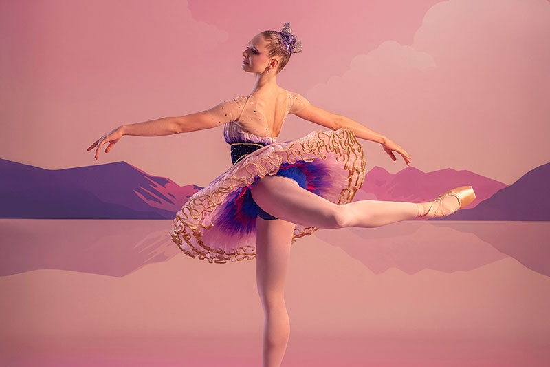 A ballerina in arabesque position, wearing a pink, purple and gold sugar plum fairy costume with a tiara. The backdrop is a pink sky and purple mountains.