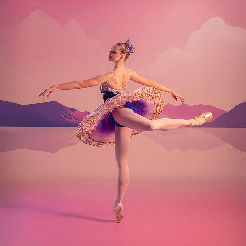 A ballerina in arabesque position, wearing a pink, purple and gold sugar plum fairy costume with a tiara. The backdrop is a pink sky and purple mountains.