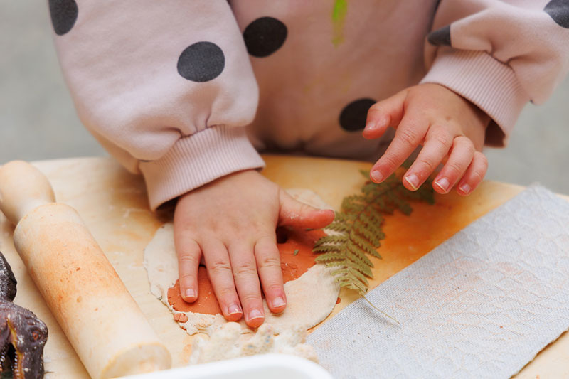 A child's hands press down a piece of clay next to a leaf and rolling pin.