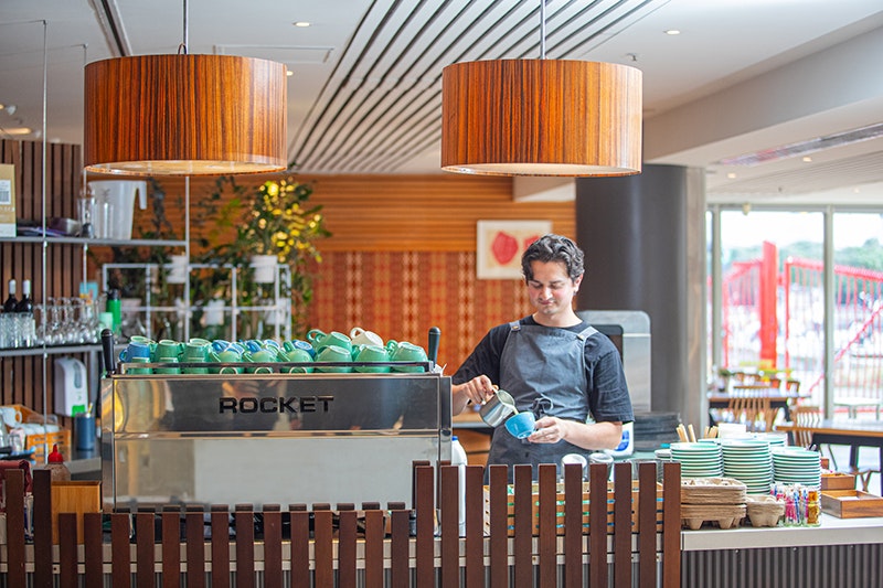 A person is standing at a coffee counter pouring milk into a cup.