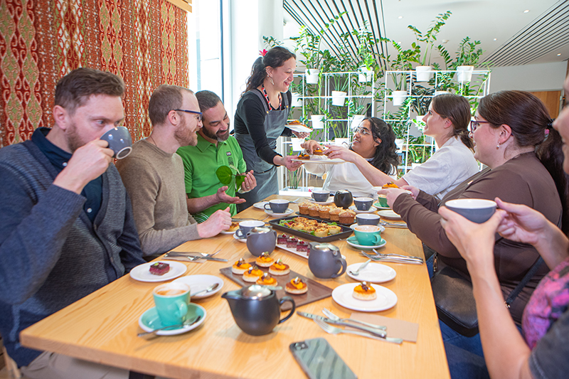 Several people are sitting around a table in a cafe. There is a server talking to some of the people and handing out a plate of food.