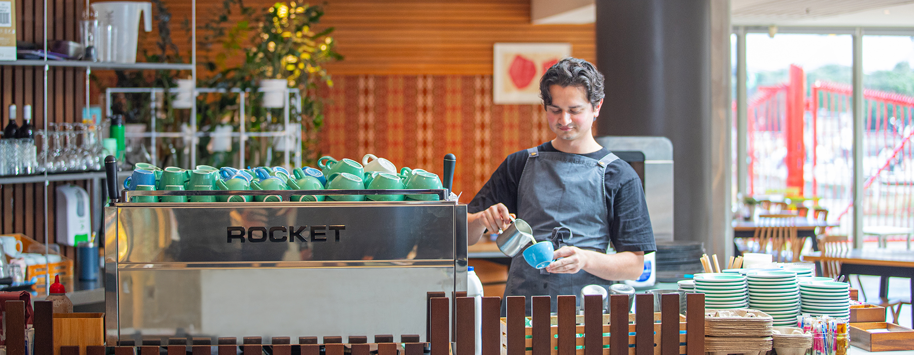 A person is standing at a coffee counter pouring milk into a cup.