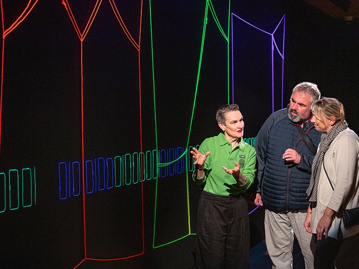 Te Papa hosts, 2025. Photo by Jeff McEwan. Te Papa (272417) A person is talking to two other people about an artwork on the wall that is brightly coloured string on a black wall.