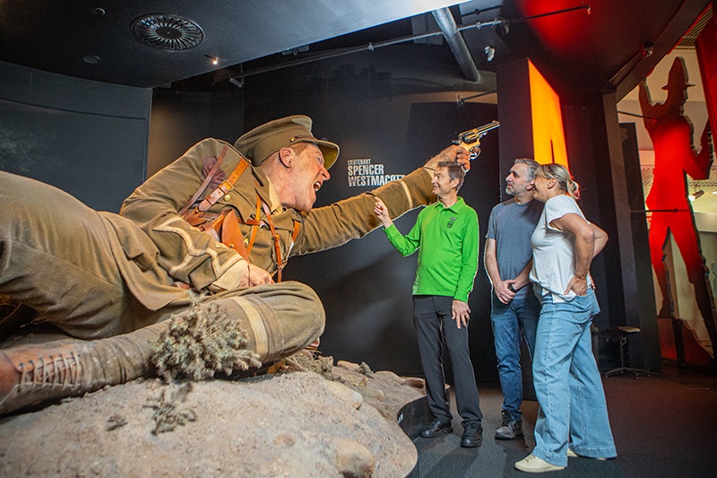 A man in a green shirt is pointing to a giant model of a soldier while talking to two other people.
