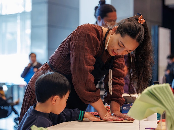 Matariki Whānau Day 2024. Photo by Jo Moore Photography. Te Papa (246256) A woman is helping a young child make a print at a large table.