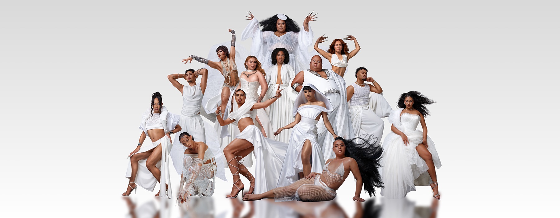 14 people dressed in glamourous style and in white are posing in a group. Some are standing, some are crouched, one is sitting on the ground.