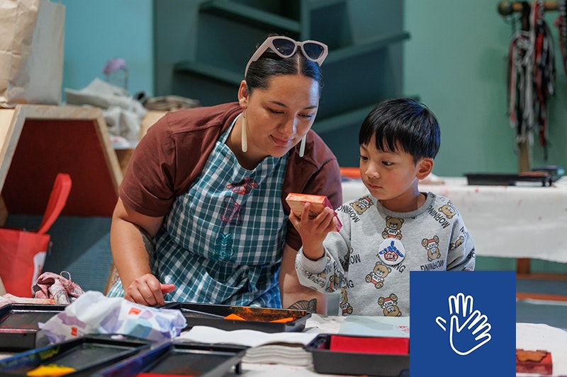 A woman with dark hair, wearning an apron sits next to a young boy with straight black hair and a grey sweatshirt. They sit side by side and look closely at the rubber stamp the boy is holding up. Ink trays and printed fabric are on the table infront. 