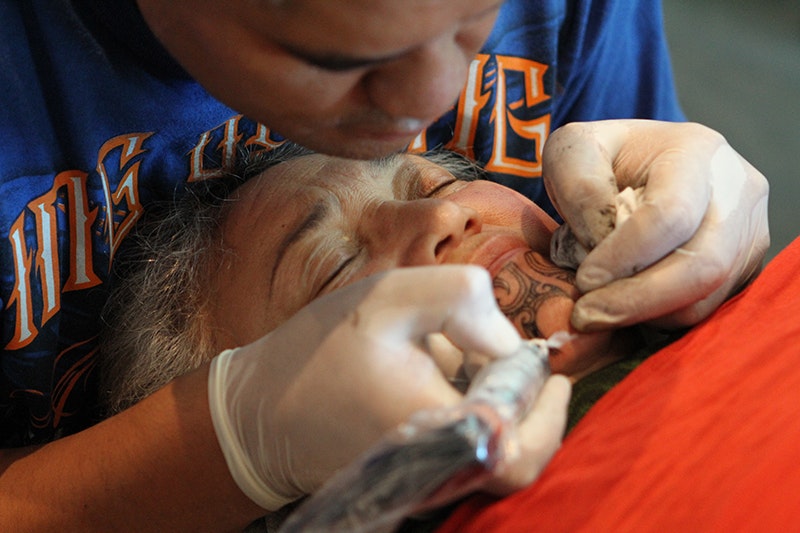 An older woman is getting a tattoo on her chin by someone with gloves on and a tattoo machine.
