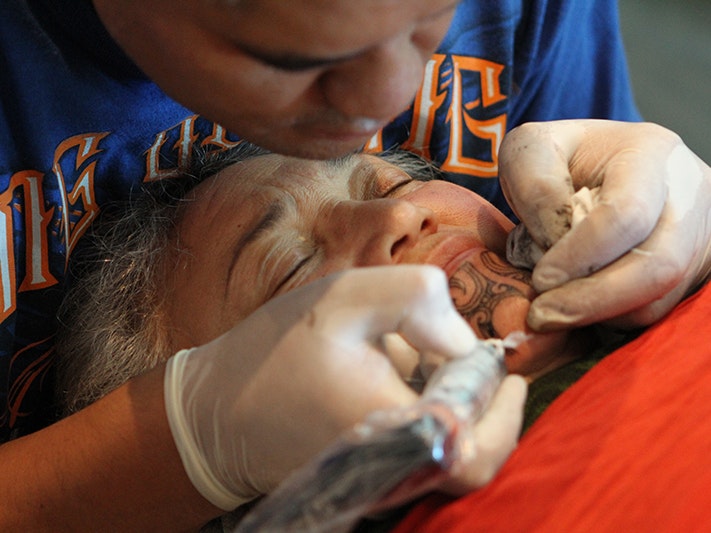 Moko on Rongomaraeroa, 2014. Te Papa (29212) An older woman is getting a tattoo on her chin by someone with gloves on and a tattoo machine.