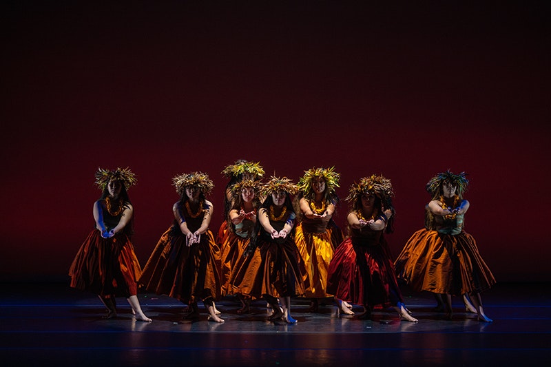 Young women performing hula on a stage, dressed in traditional Hawaiian costume. The lighting is shadowy so their faces are not completely visible. The colours the women are wearing include yellow, red, burnt orange and green. 