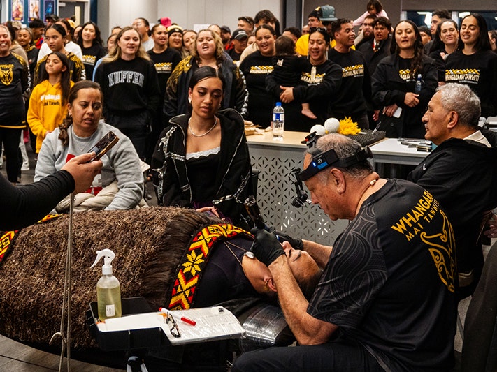 Professor Ahorangi Sir Derek Lardelli. Photo courtesy of the artist. A person wearing a headband torch is tattooing the chin of a woman lying on a table under a feather cloak. They are surrounded by a lot of people.