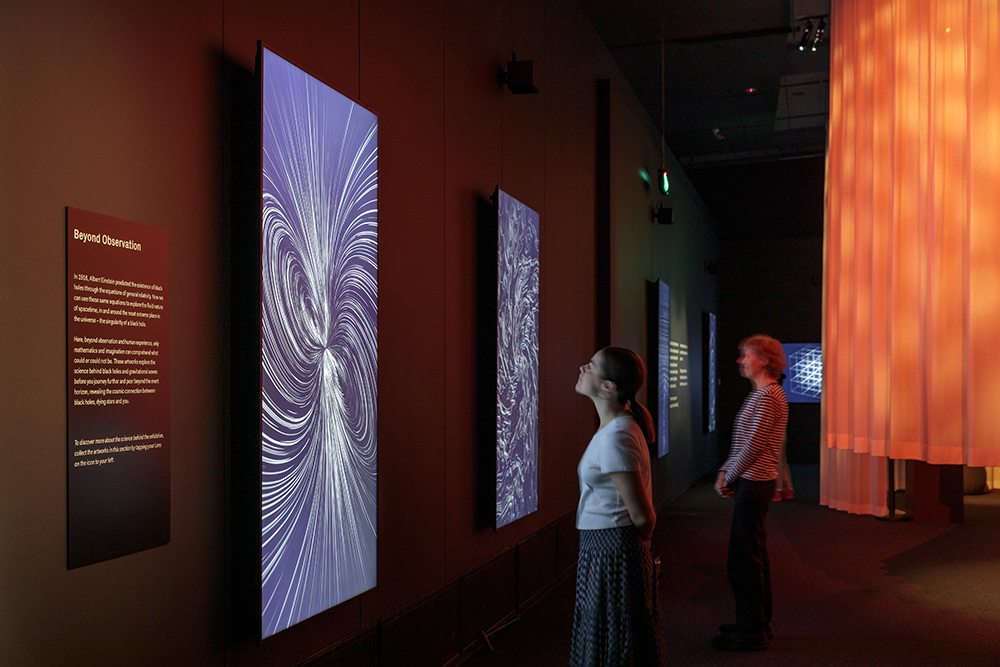 Two people are in a gallery and looking at large vertical screens on the wall that have swirling shapes on them.