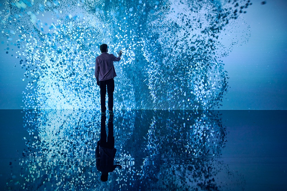A person is standing in front of a giant screen that has abstract light patterns on it. They have their hand out to it and it is all reflected on a shiny floor.