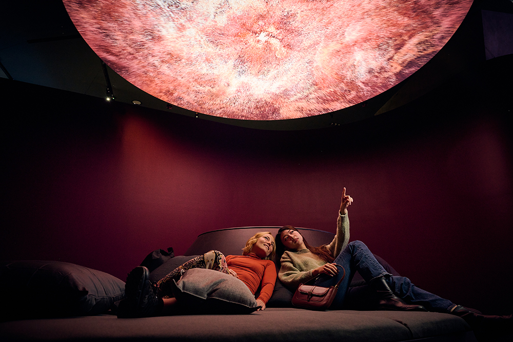 Two people are reclining on a large soft surface and looking up at a round red video above them. One of the people is pointing at it.