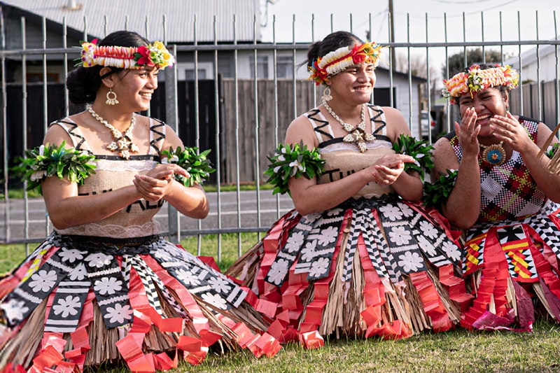 Three women in Tuvalu costume are kneeling on the grass and laughing.