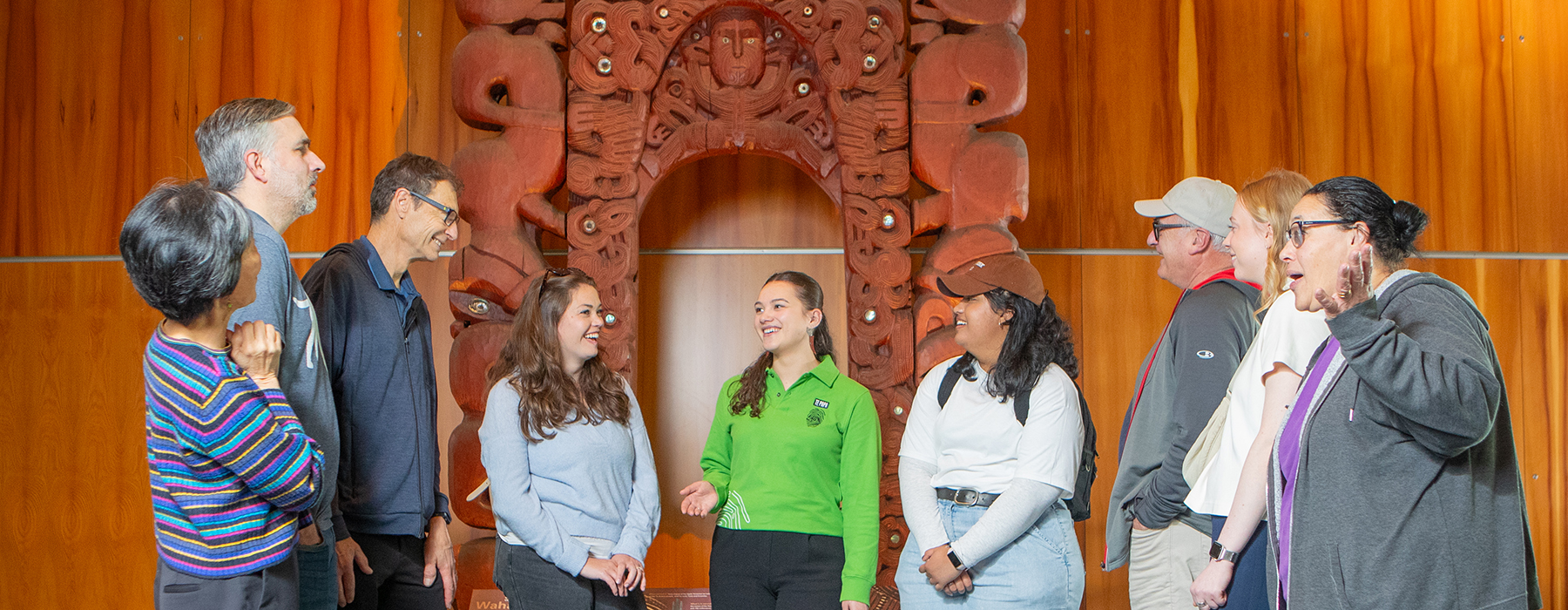 Eight people are standing either side of a woman in a bright green shirt who is talking about the wooden carving behind her.