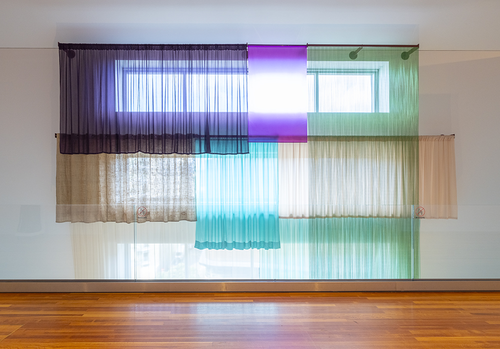 A view from a balcony of a two-floor space with three horizontal window that have several different curtains on each that are all different materials and colours.