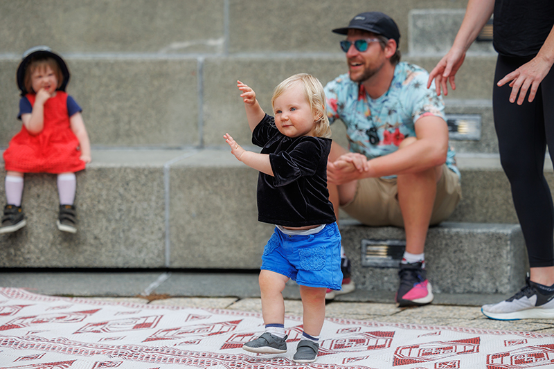 A small toddler is dancing on a mat while another toddler and two adults look on.