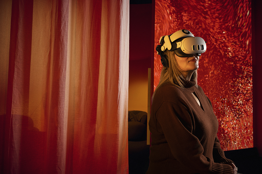 A woman with a VR headset on is standing infront of a red swirly screen and an orange curtain.