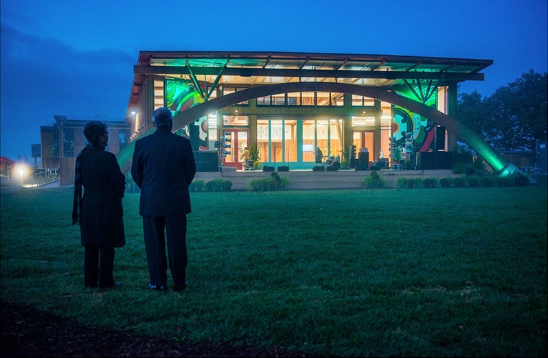 Two people with their backs to the camera are looking at a lit-up building in a grass field at twighlight.