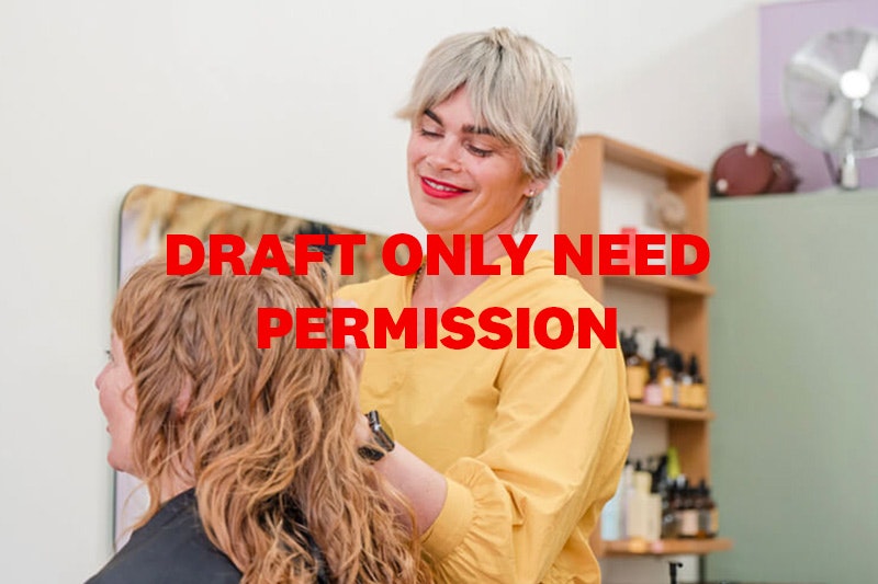 A smiling person in a yellow blouse with short blonde hair, dark eyebrows and red lipstick. She is creating waves in a clients hair.