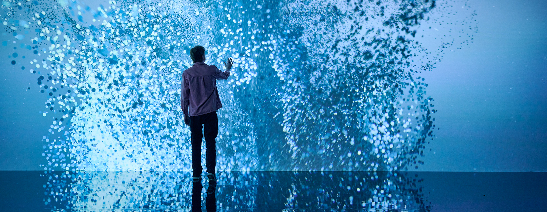 A person is standing in front of a giant screen that has abstract light patterns on it. They have their hand out to it and it is all reflected on a shiny floor.