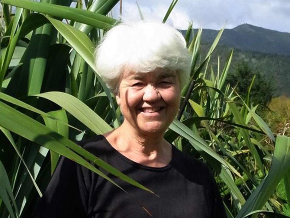 Image from Aho Kura Huna. Te Papa A woman with white hair is standing in front of a large flax bush on a sunny day.