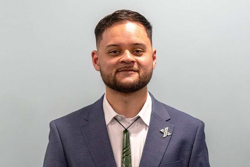 A head and shoulders photo of a man in a suit jacket and wearing a pounamu (greenstone) around his neck.