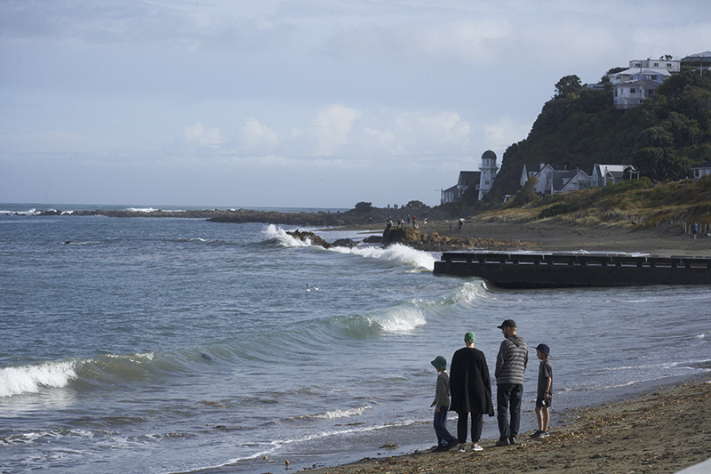 Two adults and two children are walking along a beach. There are some houses perched on a hill in the background.